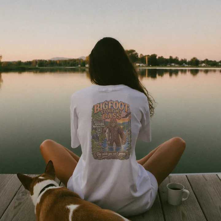 A woman sitting on the end of a lake dock beside her dog, wearing the Bigfoot Stole My Bass shirt.