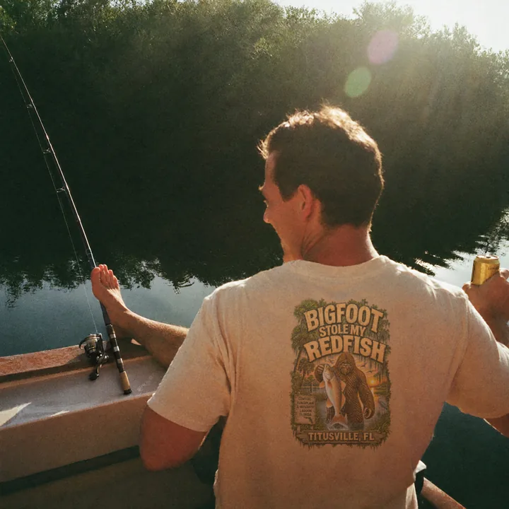 An angler with his feet up on a boat by the mangroves, beer in hand, wearing the Bigfoot Stole My Redfish shirt.