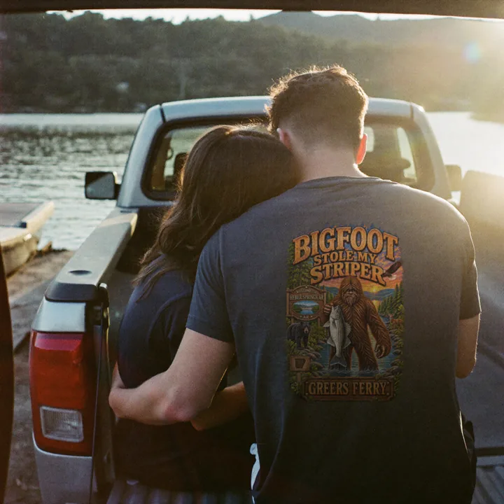 A couple on a pickup tailgate at a lake ramp at sunset, wearing the Bigfoot Stole My Striper shirt.